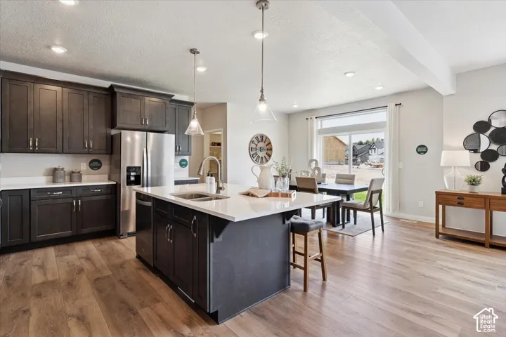 Open-concept kitchen featuring wood-finish flooring, dark wood cabinetry, stainless steel appliances, and a central island with a sink and pendant lighting
