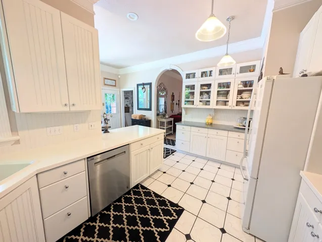 Kitchen featuring white beadboard cabinetry, solid surface countertops, stainless steel dishwasher, glass-front upper cabinets, and a black and white tiled floor