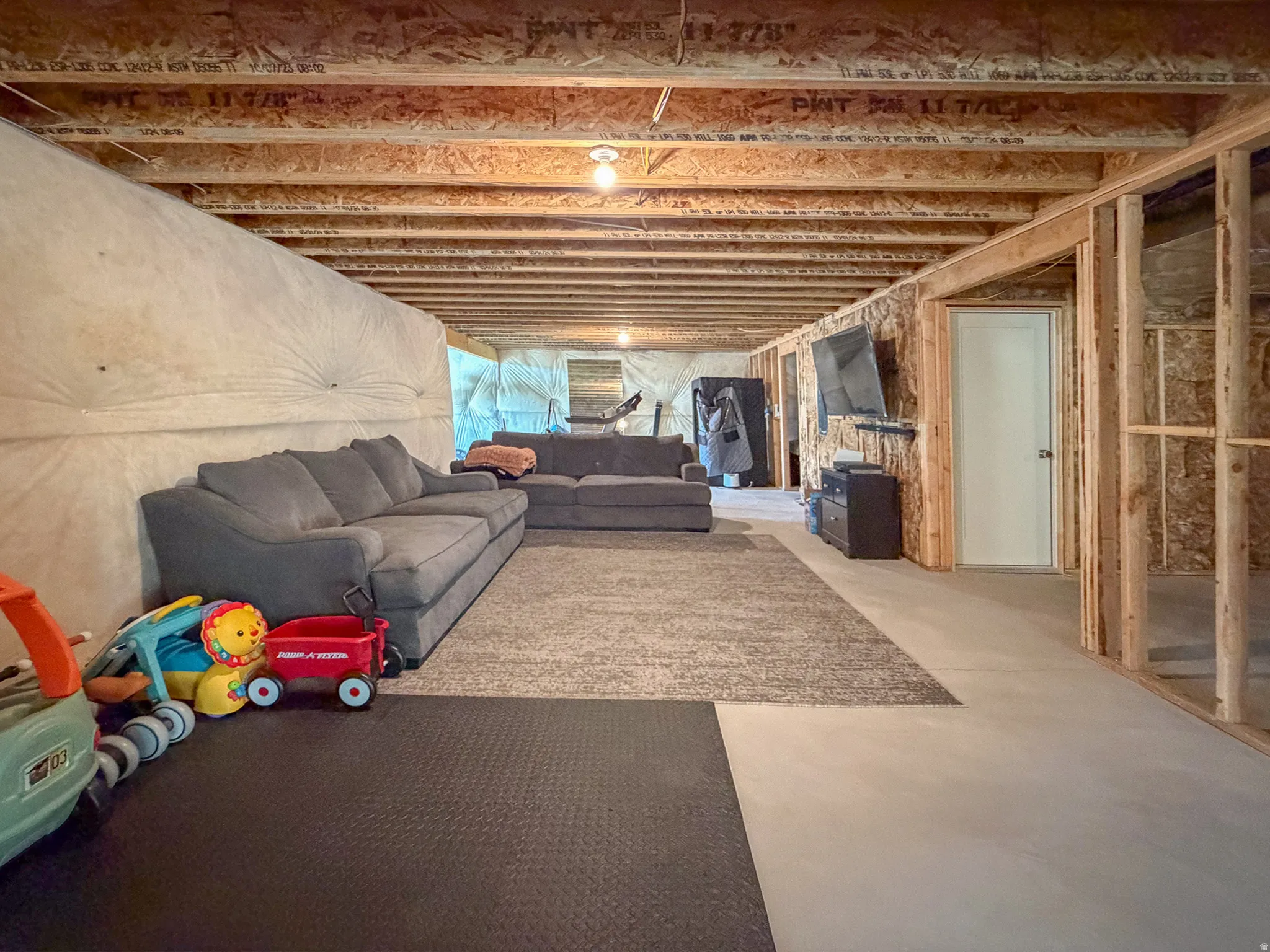 Unfinished basement with exposed ceiling joists and wood framing, featuring concrete flooring, a white interior door, and overhead lighting fixtures