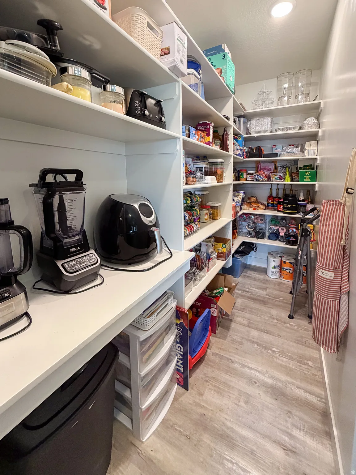 Spacious pantry featuring extensive built-in shelving, overhead lighting, durable wood-finish flooring, and ample counter space