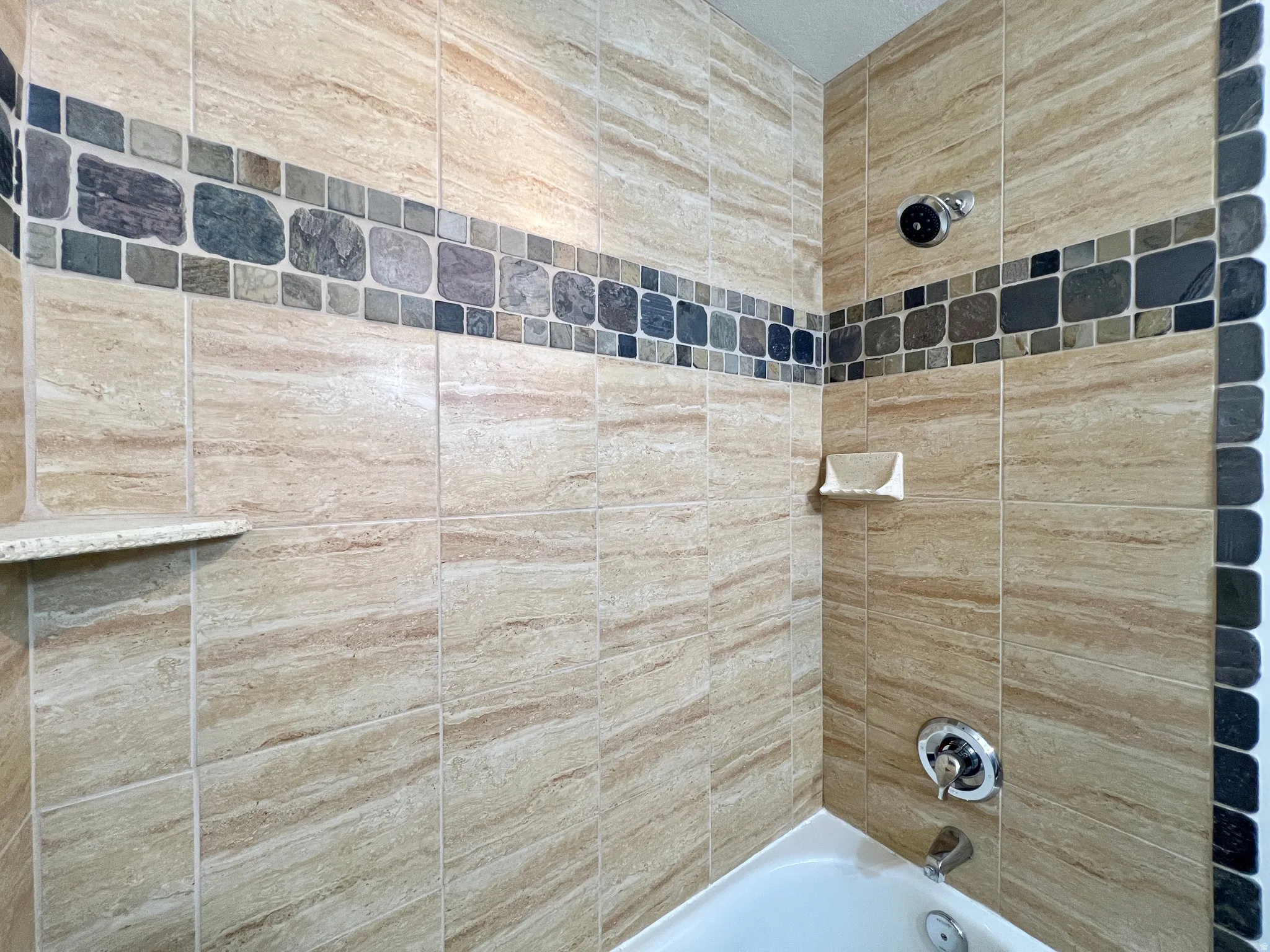 Shower featuring floor-to-ceiling ceramic tile, a decorative stone mosaic accent band, and a built-in corner shelf