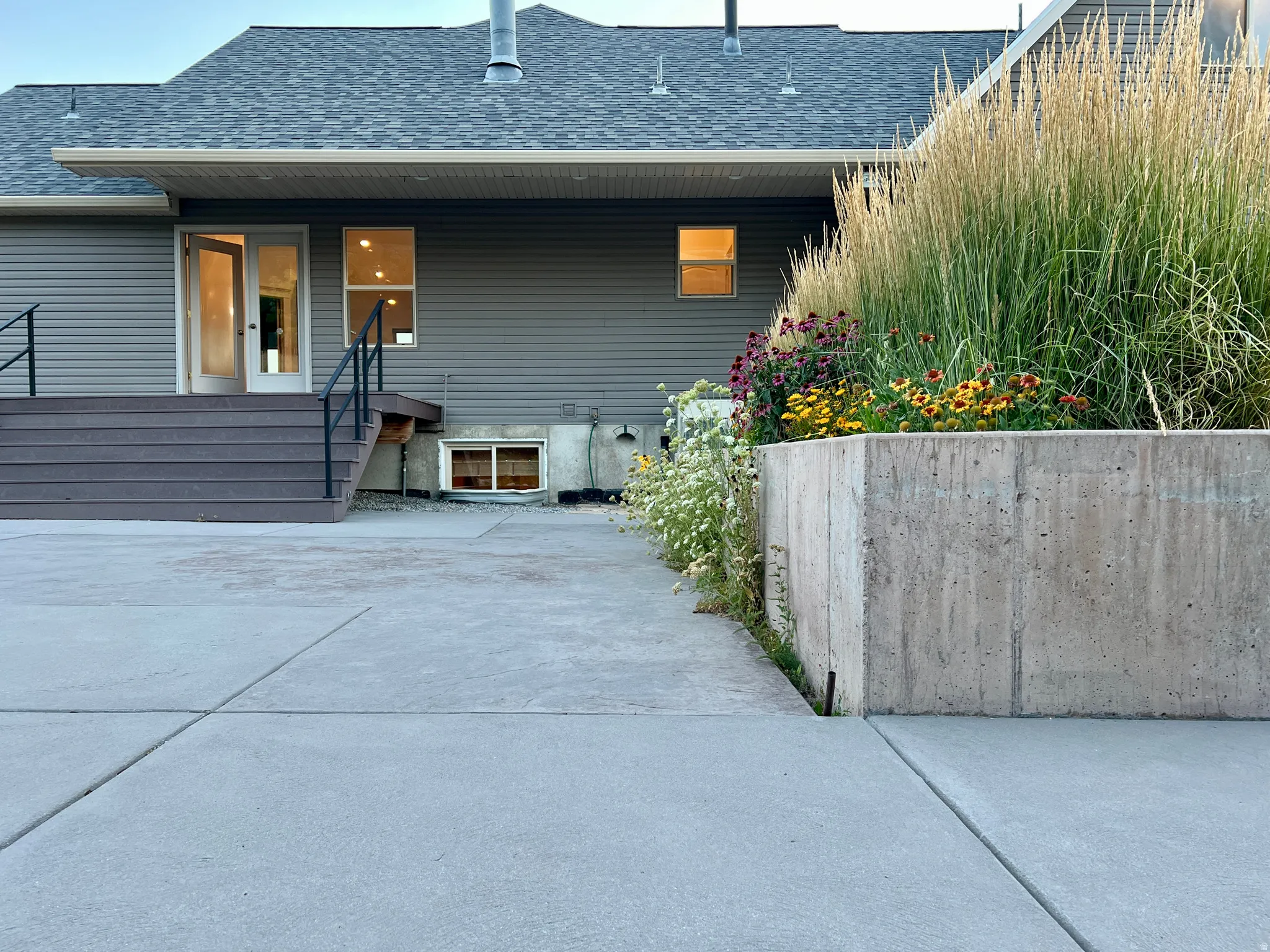 Exterior concrete patio with a concrete retaining wall, composite deck stairs with black railings, and a planted garden bed featuring ornamental grasses and various flowering plants