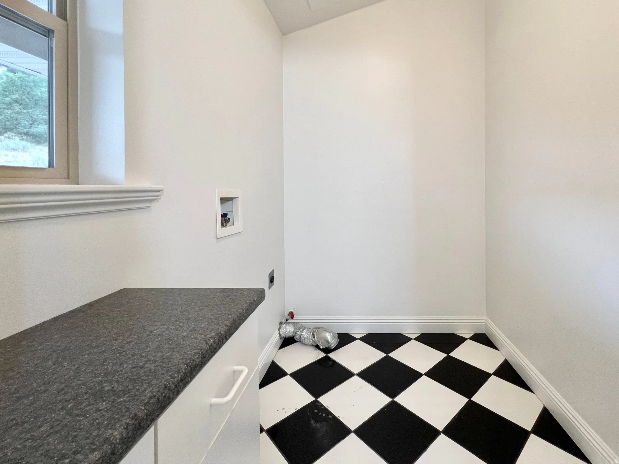 Utility space with checkerboard flooring, a window providing natural light, a dark speckled countertop, and white base cabinetry with a single drawer