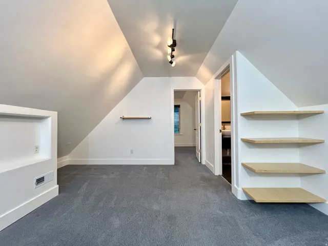 Carpeted space with sloped ceilings, track lighting, built-in shelving, and a recessed wall niche with an electrical outlet