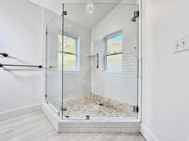 Glass-enclosed shower featuring white subway tile walls, a pebble-stone floor, and two windows