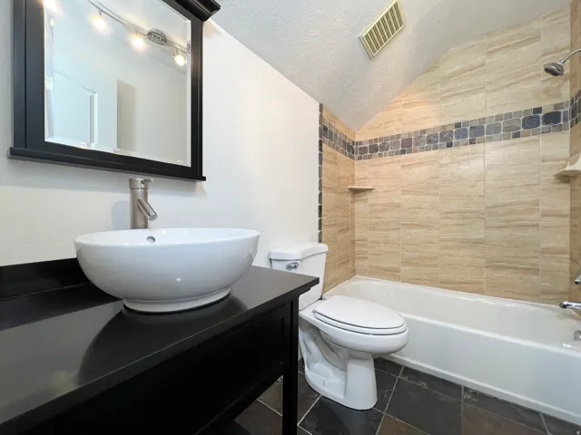 Bathroom featuring a white vessel sink, black vanity with a matching framed mirror, brushed nickel faucet, and a tiled shower/tub combination with an accent band and built-in shelving
