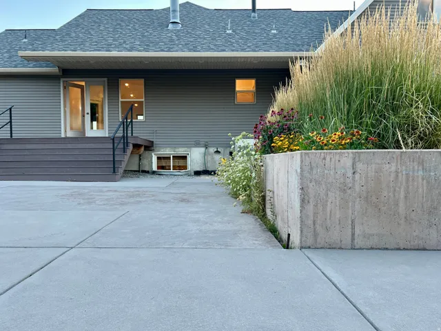 Exterior concrete patio with a concrete retaining wall, composite deck stairs with black railings, and a planted garden bed featuring ornamental grasses and various flowering plants