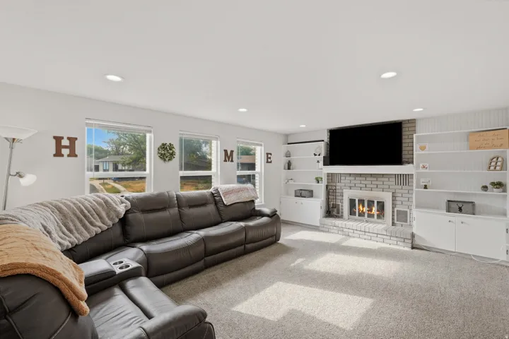 Spacious living area featuring a brick-surround fireplace, built-in shelving, recessed lighting, multiple windows, and light-toned carpeting