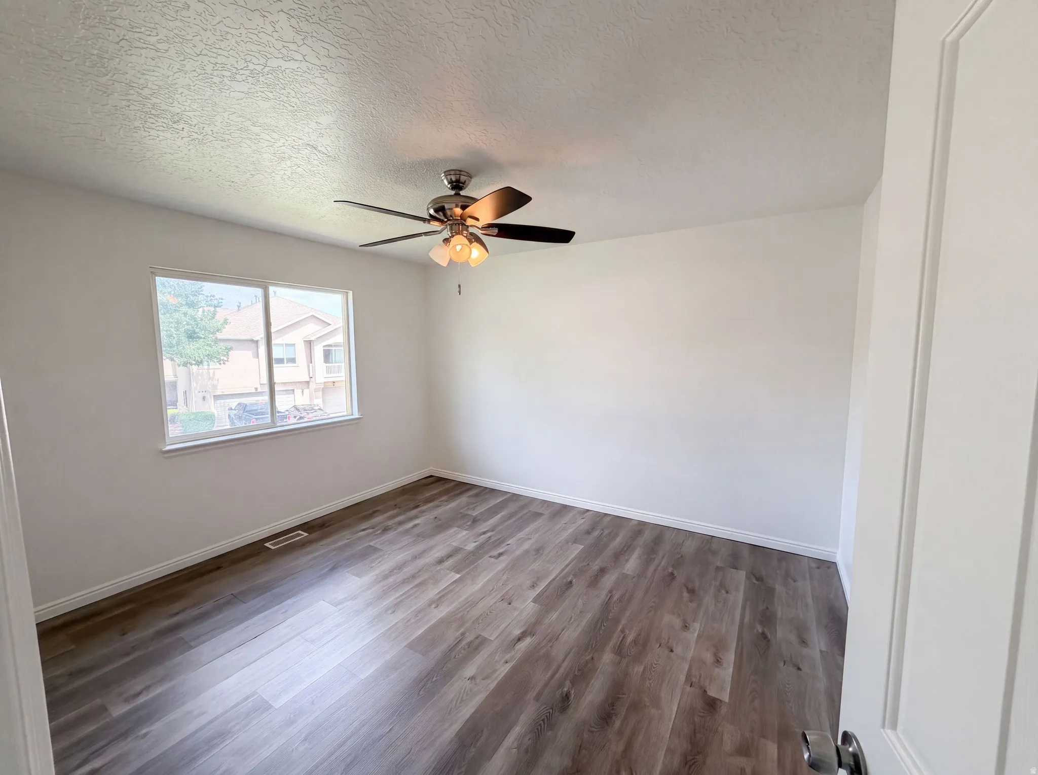 Bright interior space featuring wood-finish flooring, a contemporary ceiling fan with integrated lighting, a large window with white trim, and neutral wall tones