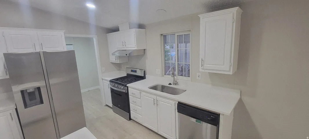 Kitchen featuring white cabinetry, light-toned countertops, a stainless steel refrigerator, a gas range with an overhead vent, and a stainless steel sink with a window above