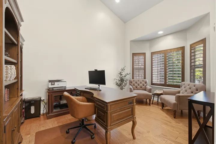 Sophisticated home office featuring a wood-finish desk with turned legs, warm wood cabinetry with open shelving, hardwood flooring, recessed ceiling lighting, and wooden shuttered windows providing natural light and privacy control