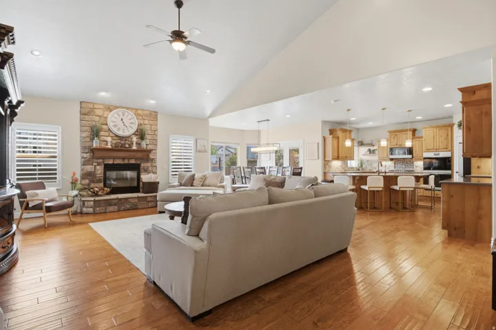 Vaulted ceiling with a ceiling fan, stone-faced fireplace with a wood mantle, wood-finish flooring, and white plantation shutters