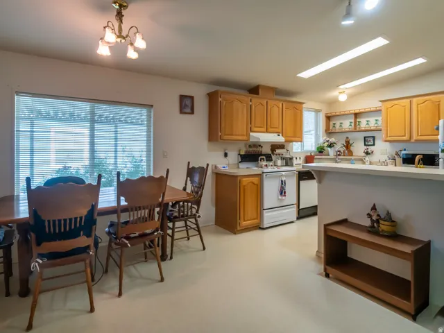 Open-concept kitchen and dining area featuring light wood cabinetry, white appliances, a breakfast bar, and a large dining room window with horizontal blinds