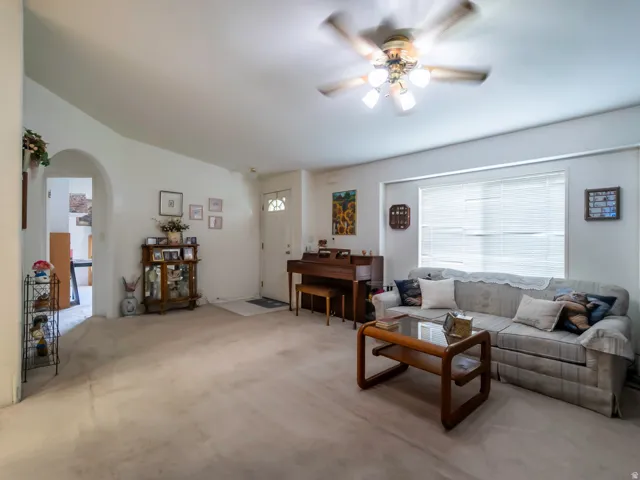 Living area featuring neutral-toned carpeting, a ceiling fan with integrated lighting, and a large window with horizontal blinds