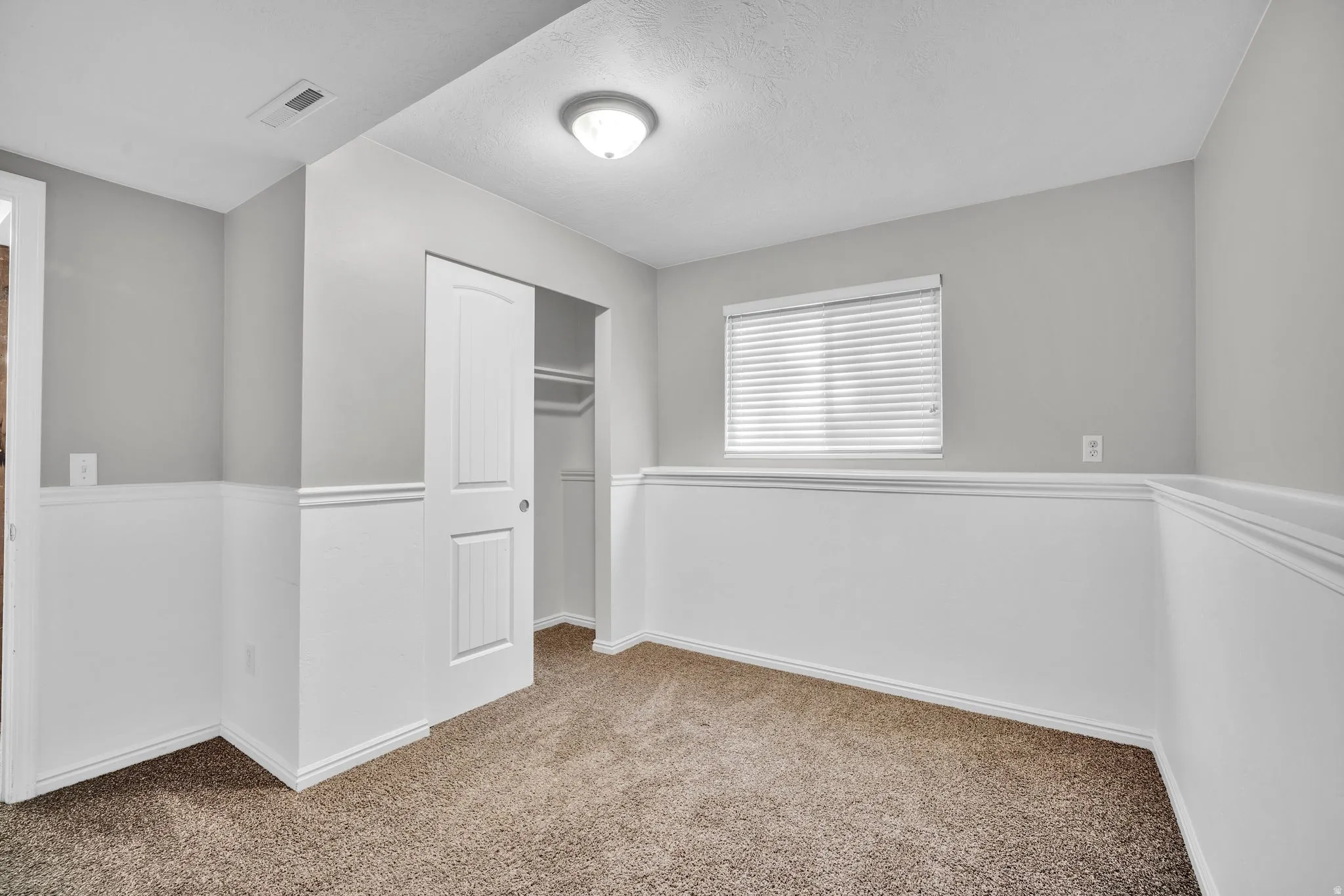 Carpeted room with neutral wall paint and white wainscoting, featuring a window with horizontal blinds, a ceiling-mounted light fixture, and a sliding closet door