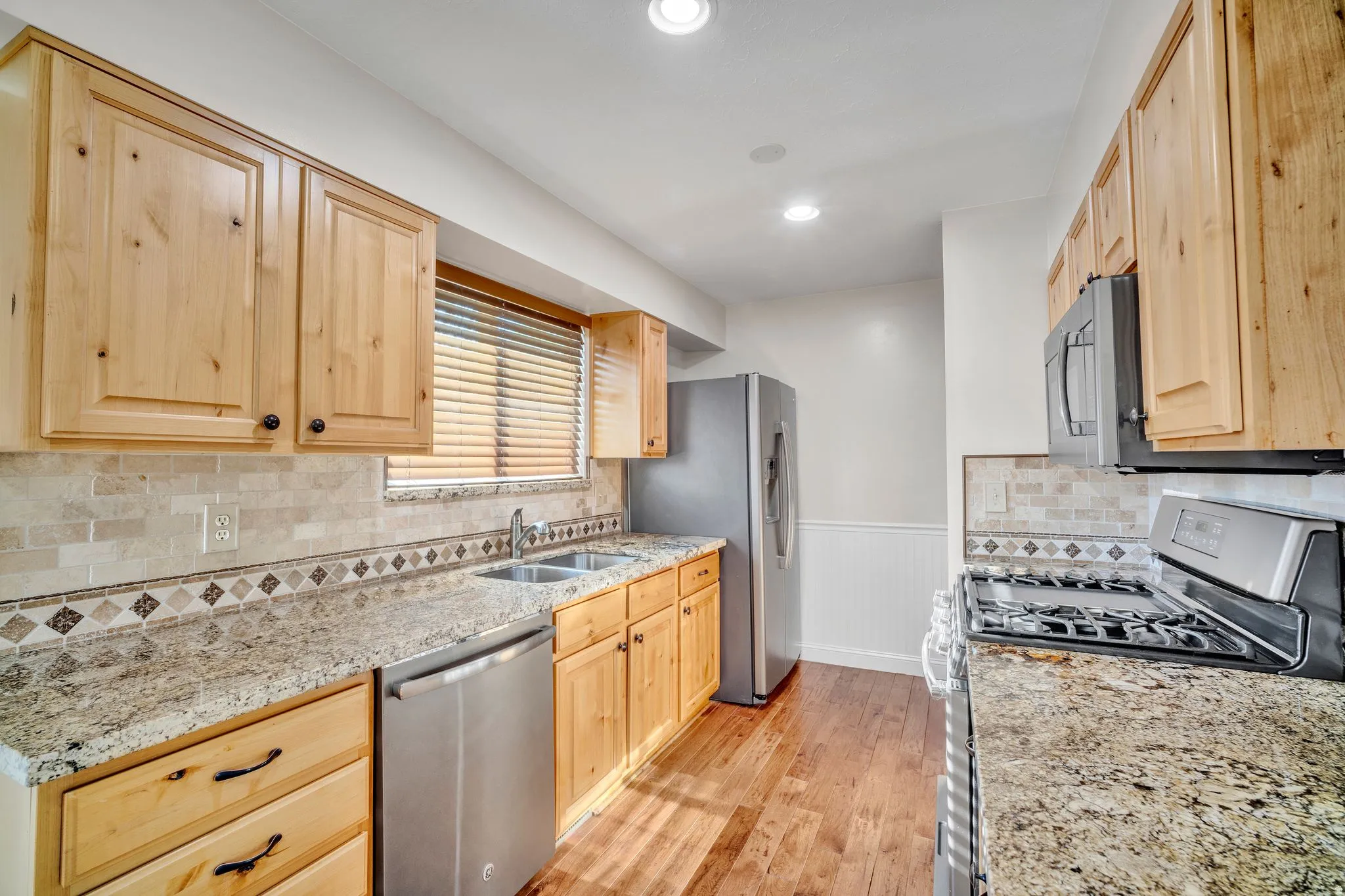Kitchen featuring wood-finish cabinetry, granite-style countertops, stainless steel appliances, a tile backsplash with a decorative border, and wood-finish flooring