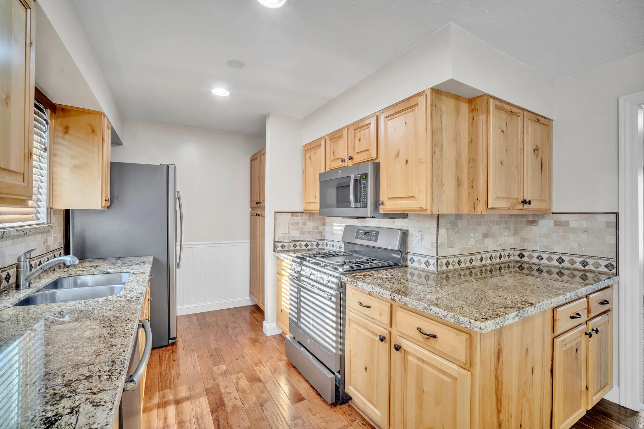 Kitchen featuring wood-finish flooring, light wood cabinetry, granite-style countertops, stainless steel appliances, and a tiled backsplash