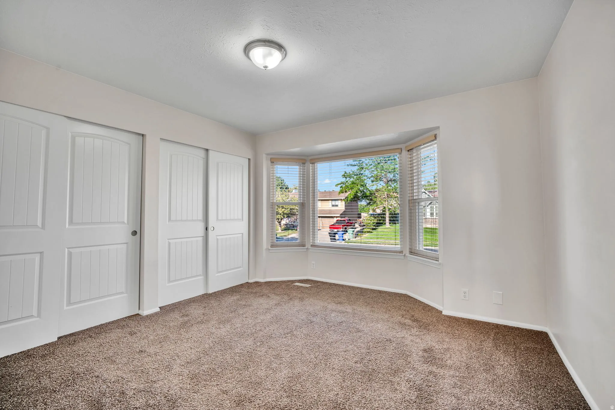 Carpeted room featuring a bay window with blinds, dual sliding closet doors, and an overhead flush-mount light fixture