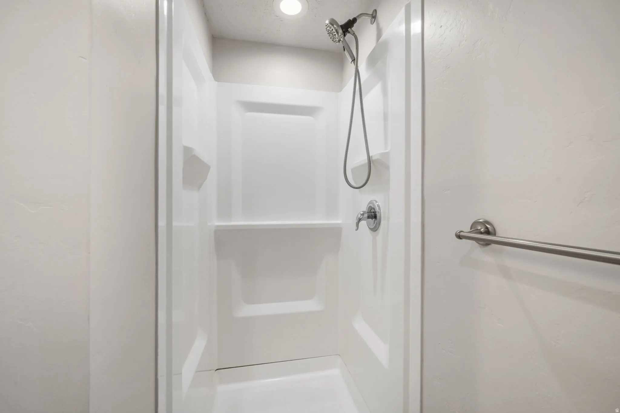 White shower enclosure featuring integrated shelving, recessed lighting, a handheld showerhead, and a towel bar