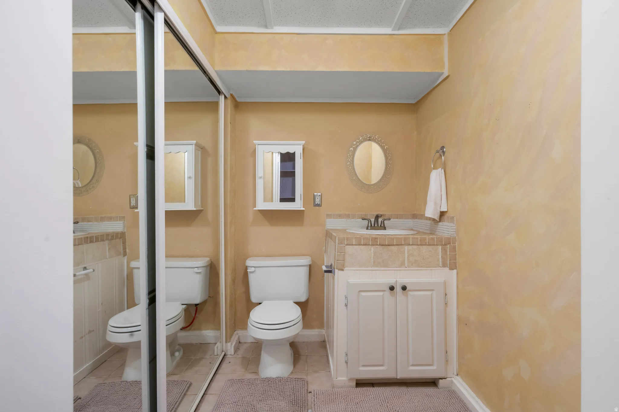 Bathroom featuring a single-sink vanity with a tiled countertop and white cabinetry, a toilet, a wall-mounted medicine cabinet, and a mirrored closet