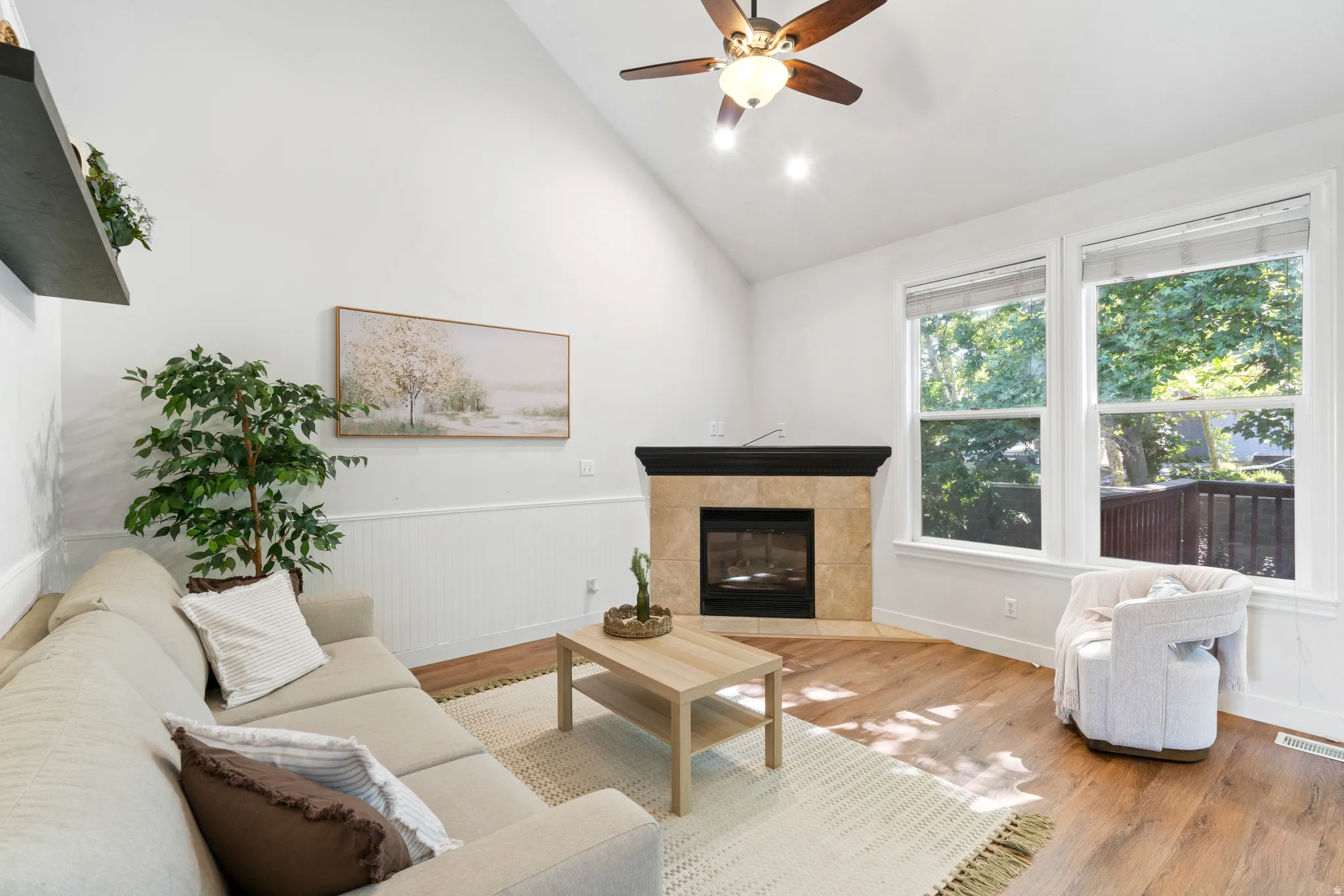 Living area featuring a vaulted ceiling with a ceiling fan, a corner fireplace with a tile surround and dark wood mantel, wood-finish flooring, and large windows with white trim