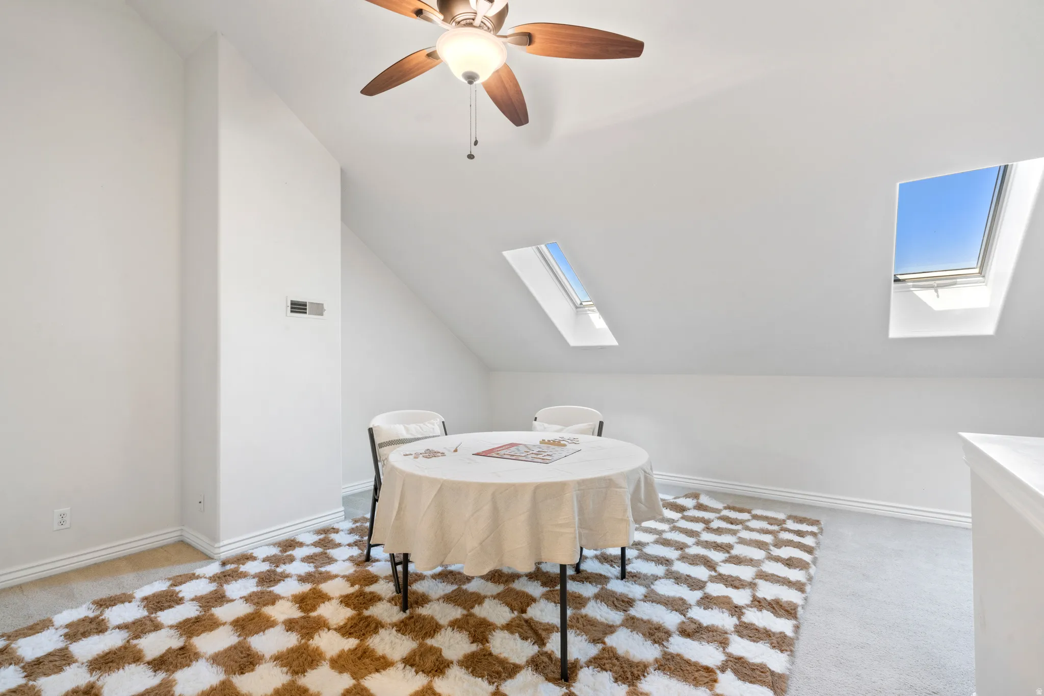 Attic space featuring two skylights, a ceiling fan with integrated lighting, white walls, and light-colored carpeting