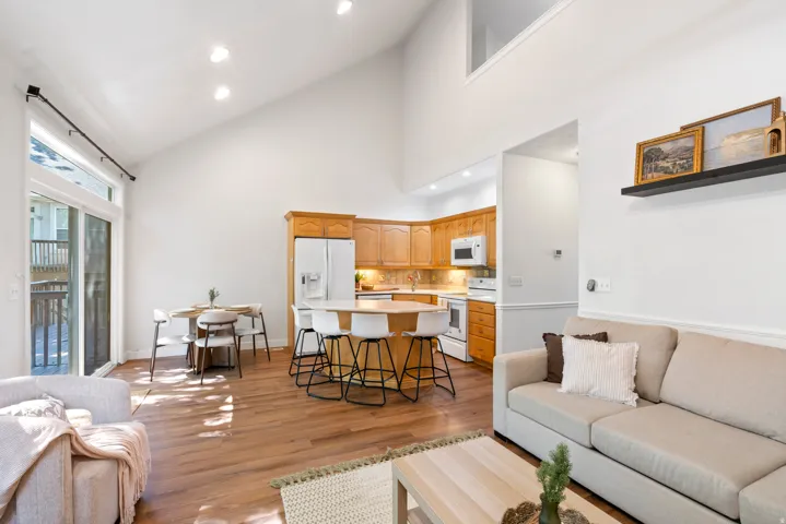 Open-concept living area featuring vaulted ceilings, wood-finish flooring, a full kitchen with light wood cabinetry, and a dining area with direct access to an outdoor deck