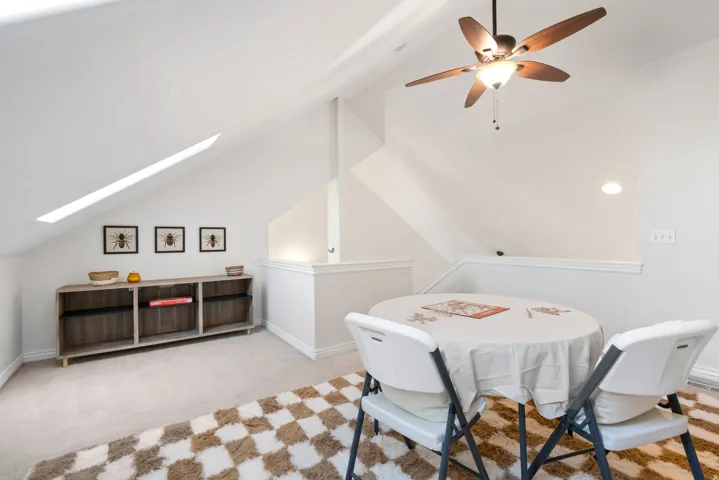 Carpeted attic space featuring a built-in shelving unit, a sloped ceiling with a skylight, and a ceiling fan with integrated lighting