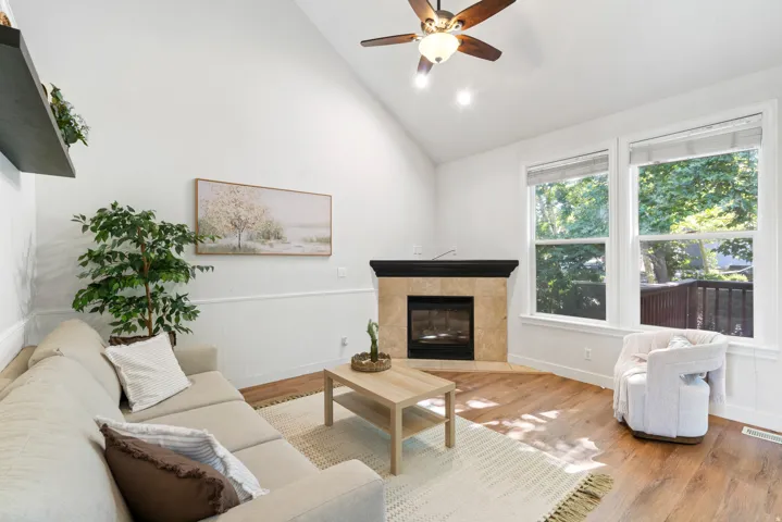 Living area featuring a vaulted ceiling with a ceiling fan, a corner fireplace with a tile surround and dark wood mantel, wood-finish flooring, and large windows with white trim