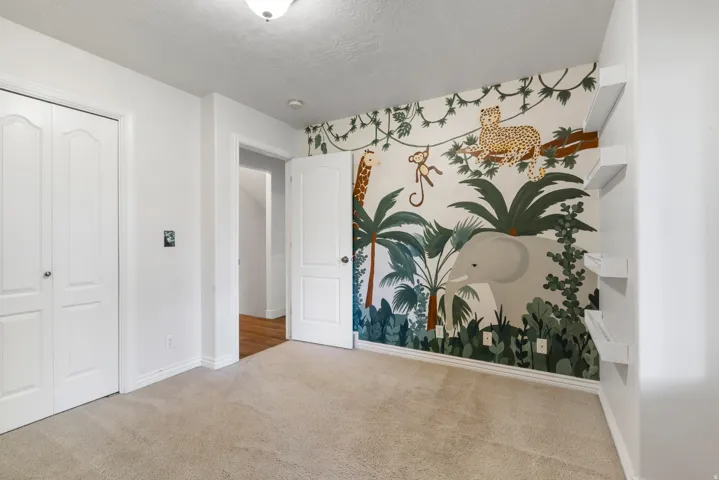 Bedroom featuring a statement accent wall with a jungle mural, light-toned carpeting, white trim, and a ceiling-mounted light fixture