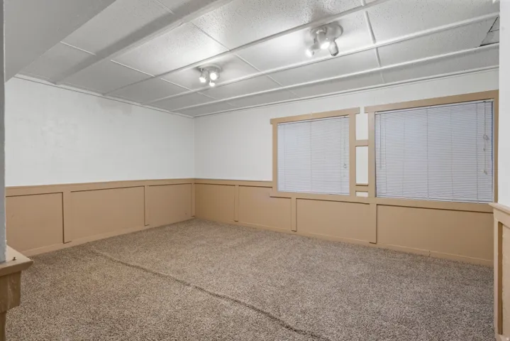 Carpeted room featuring two windows with horizontal blinds, board and batten style wainscoting, white walls, and a drop ceiling with recessed lighting