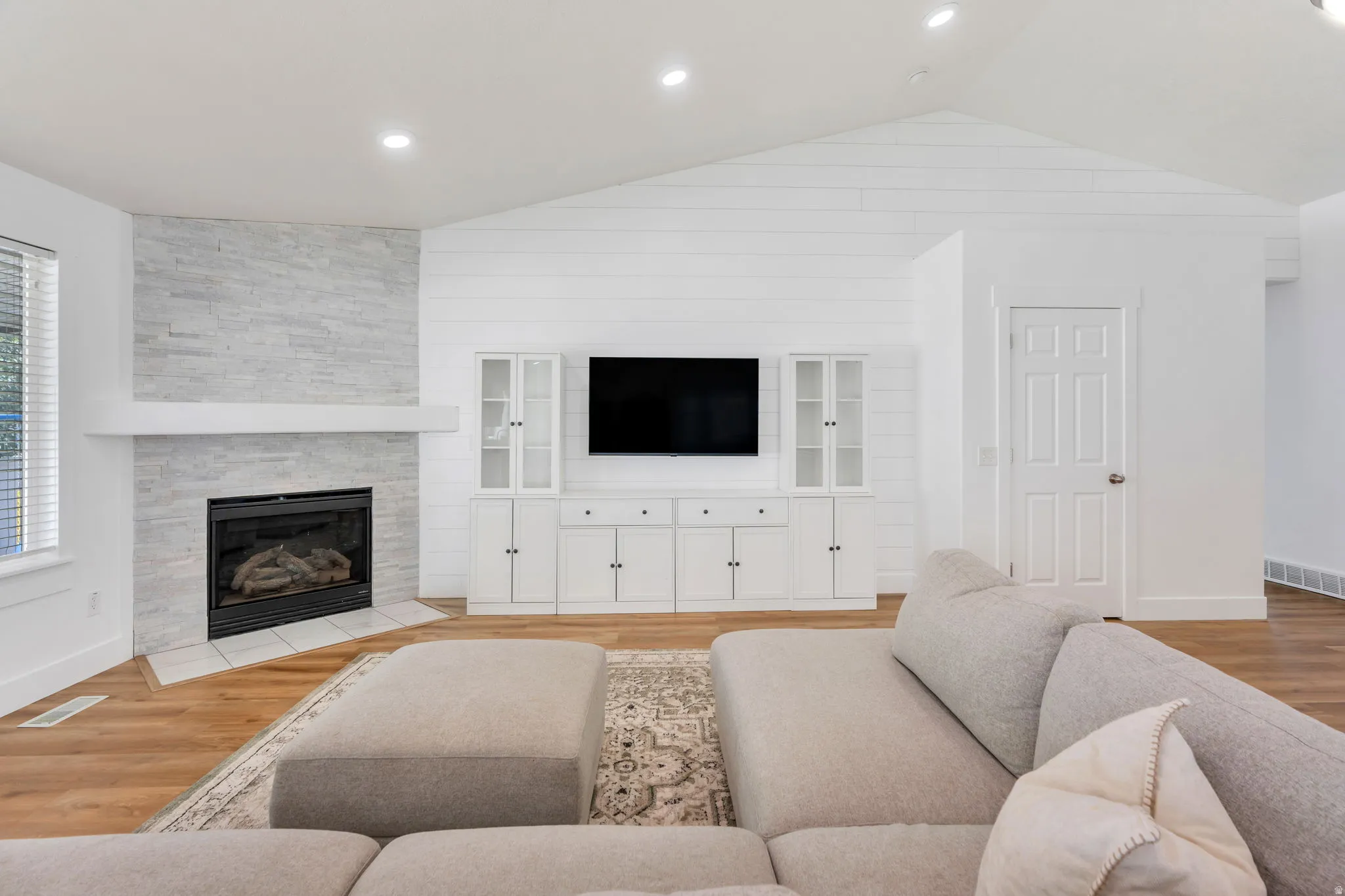 Living area featuring wood-finish flooring, a stone-clad fireplace with a hearth and white mantle, built-in entertainment cabinetry with glass-front display cabinets, and a vaulted ceiling with recessed lighting