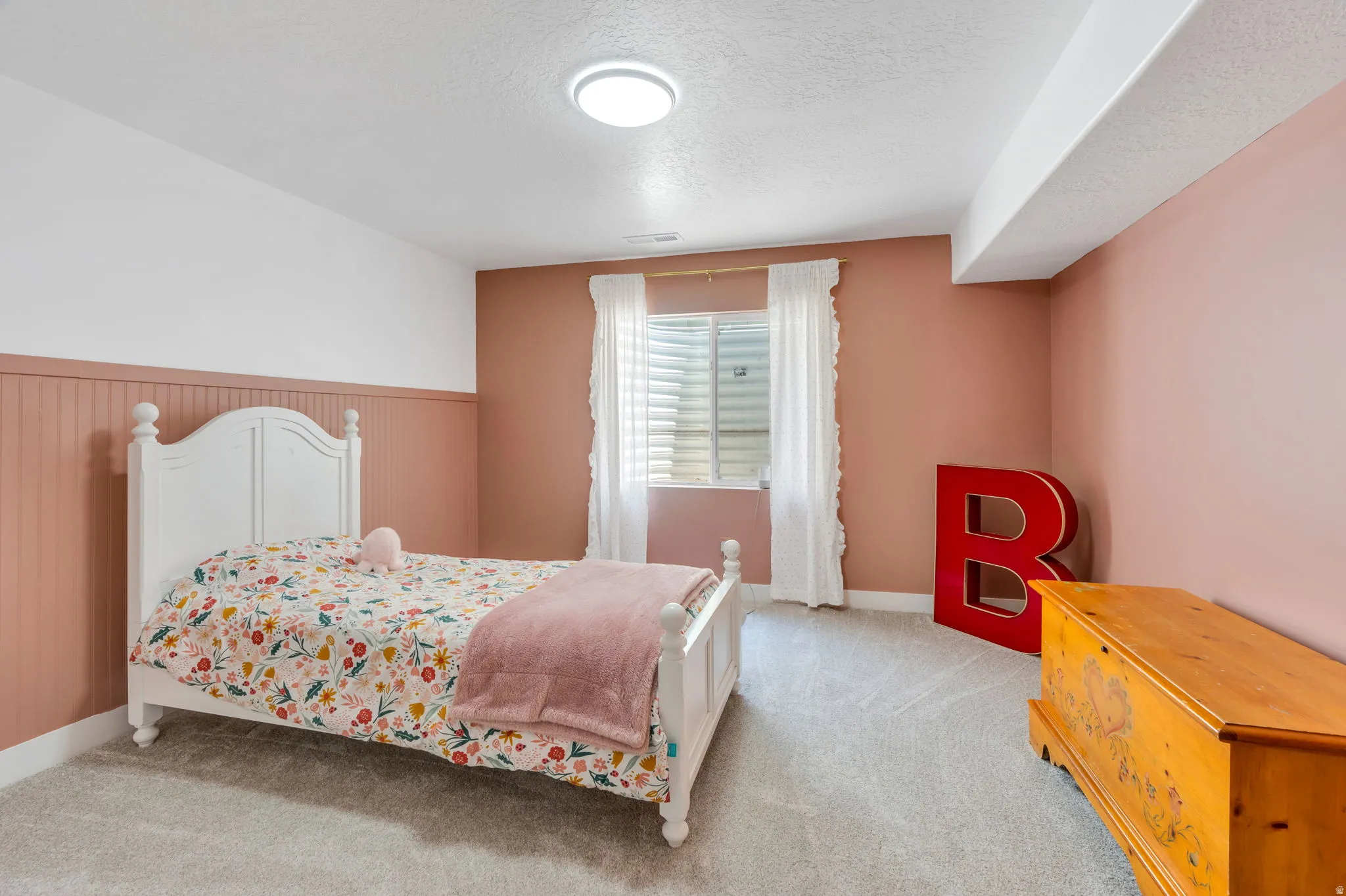 Room featuring a window with white blinds and sheer curtains, light-colored carpeting, a partial accent wall with vertical paneling, and a flush-mount ceiling light fixture