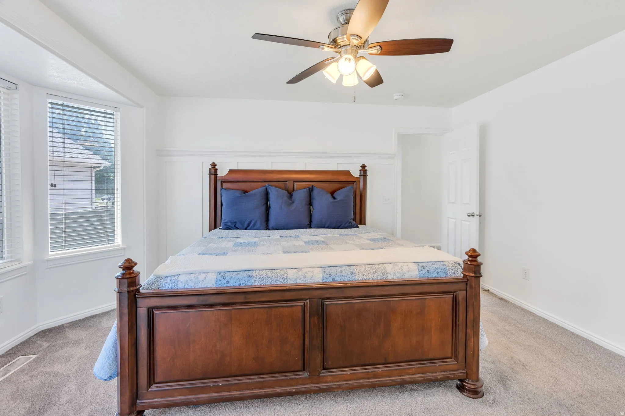 Spacious primary bedroom featuring a solid wood four-poster bed frame, neutral carpeting, ceiling fan with integrated lighting, and abundant natural light from dual windows with plantation shutters