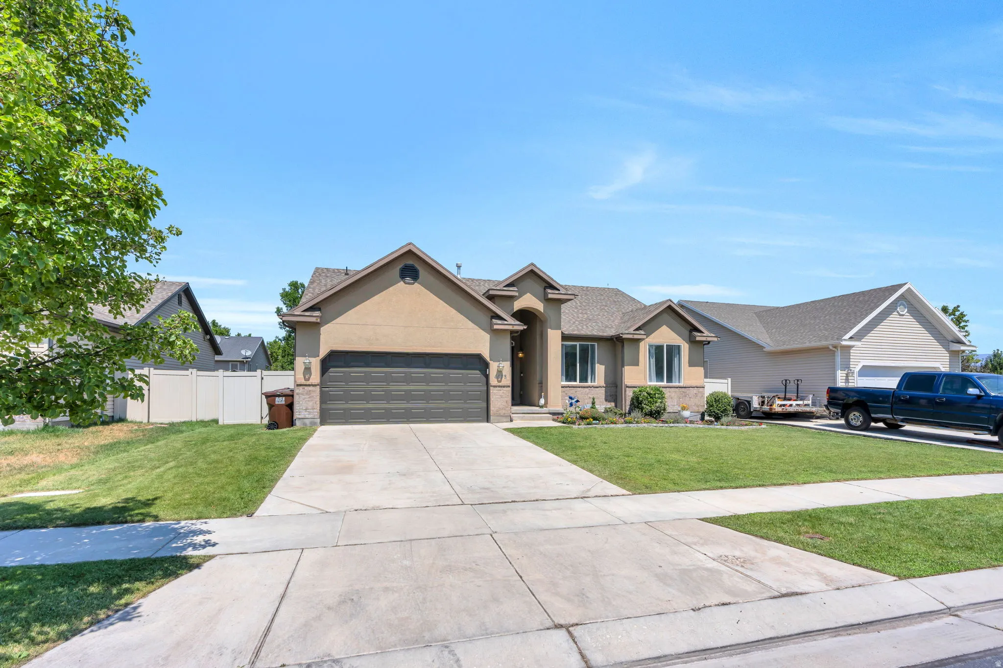 Welcoming front elevation featuring a tan stucco exterior with peaked roofline, dark-finish two-car garage, concrete driveway with geometric sections, and manicured lawn bordered by mature landscaping