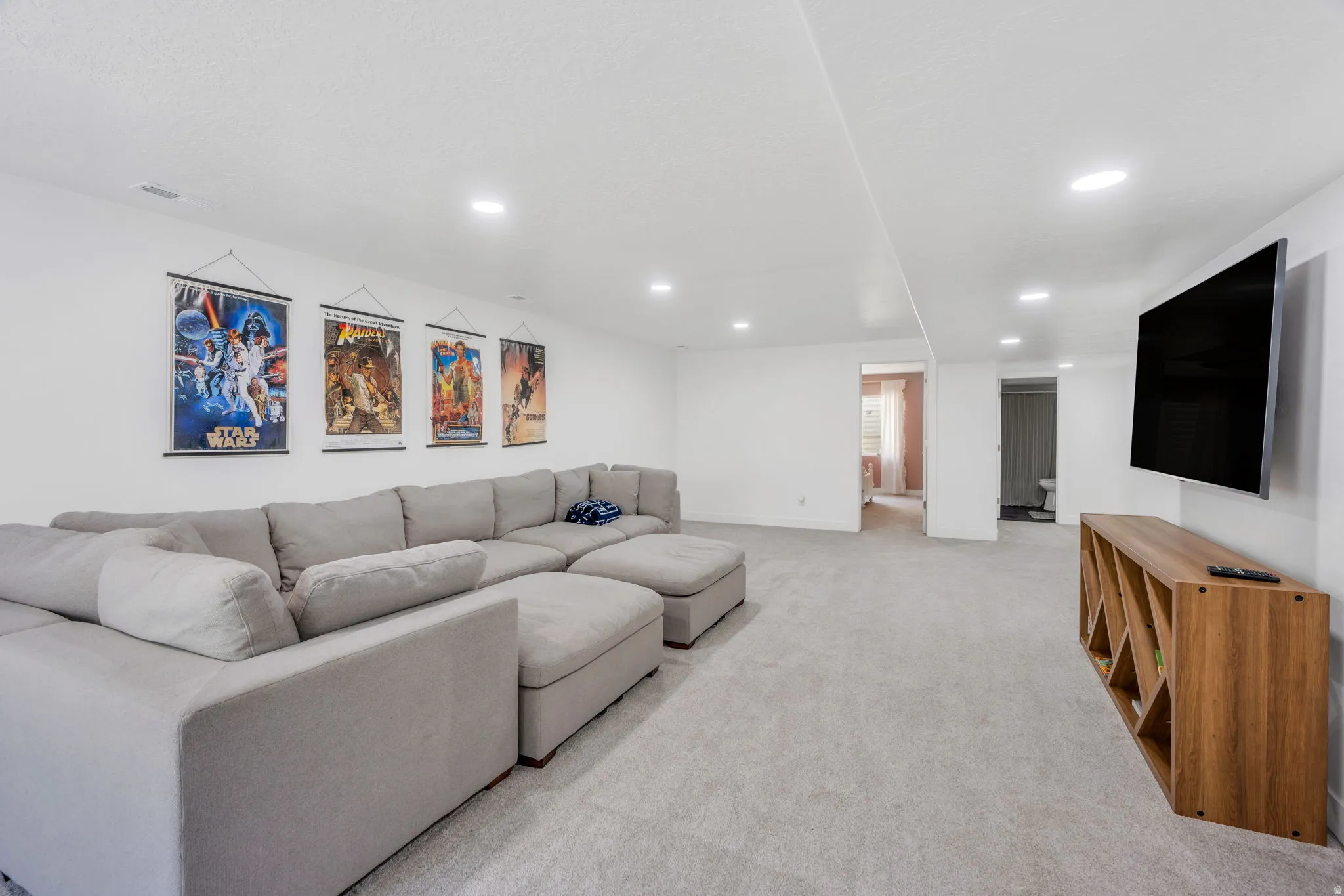 Spacious recreational room featuring a sectional seating arrangement, recessed ceiling lighting, wall-mounted flat-screen television above wood-finish media cabinetry, and neutral carpet flooring throughout