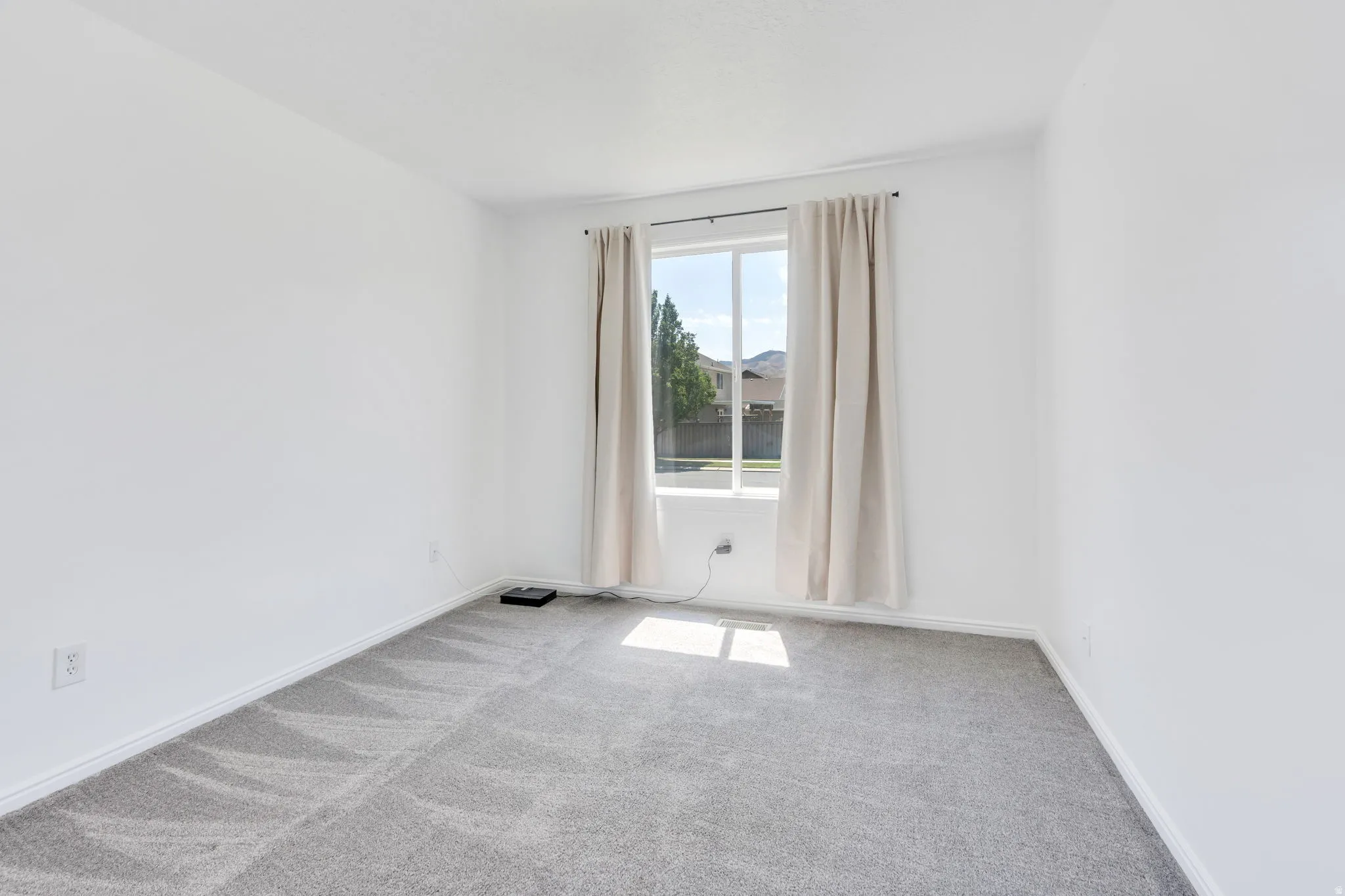 Light-filled room featuring a large window, neutral wall paint, gray carpeting, white baseboards, and a window treatment