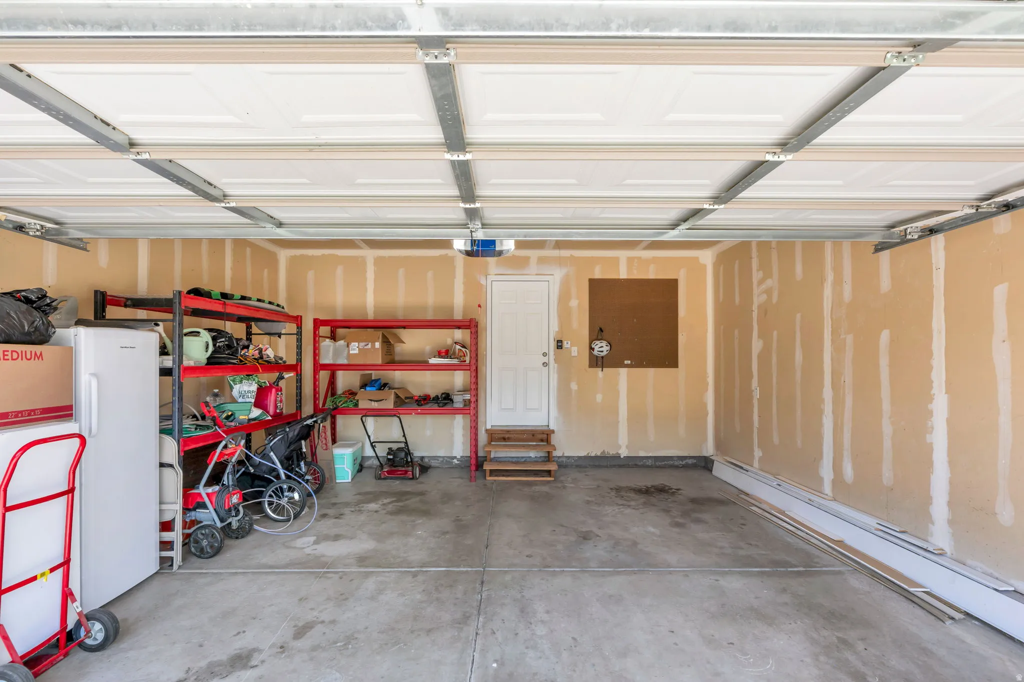 Spacious attached garage featuring epoxy-coated concrete flooring, overhead LED lighting system, built-in red metal shelving units, and white overhead garage door with natural light panels