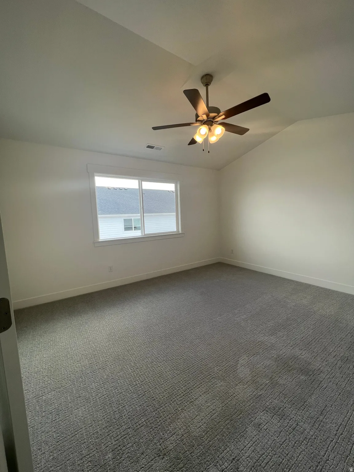 Spacious room featuring vaulted ceilings, a contemporary ceiling fan with integrated lighting, a large window, and textured gray carpeting