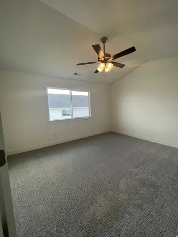 Spacious room featuring vaulted ceilings, a contemporary ceiling fan with integrated lighting, a large window, and textured gray carpeting