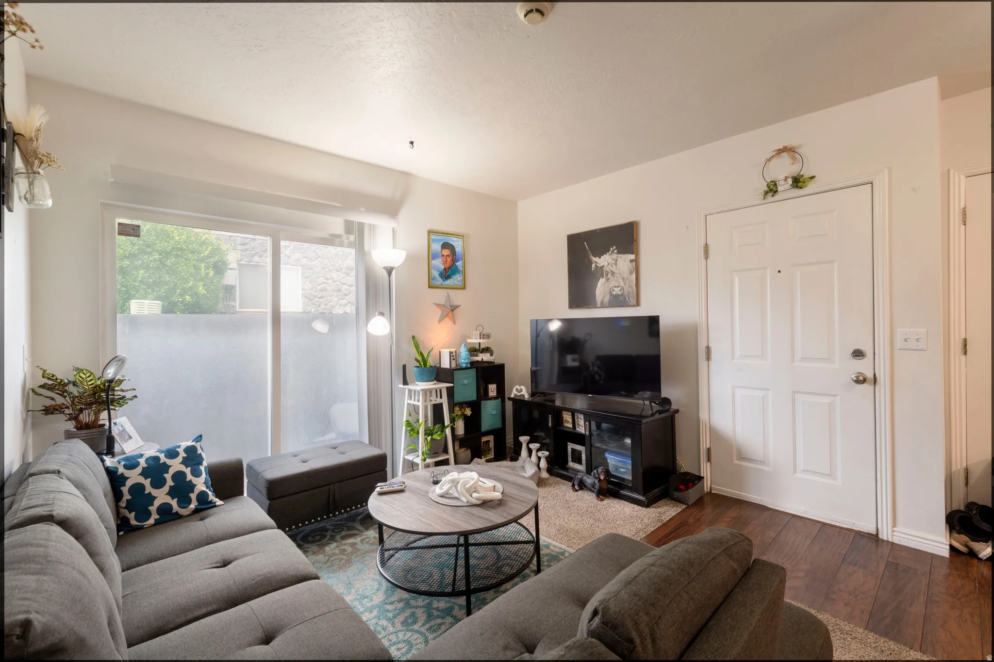 Spacious living area featuring a vaulted ceiling, wood-finish flooring, a white paneled entry door, and a sliding glass door providing natural light