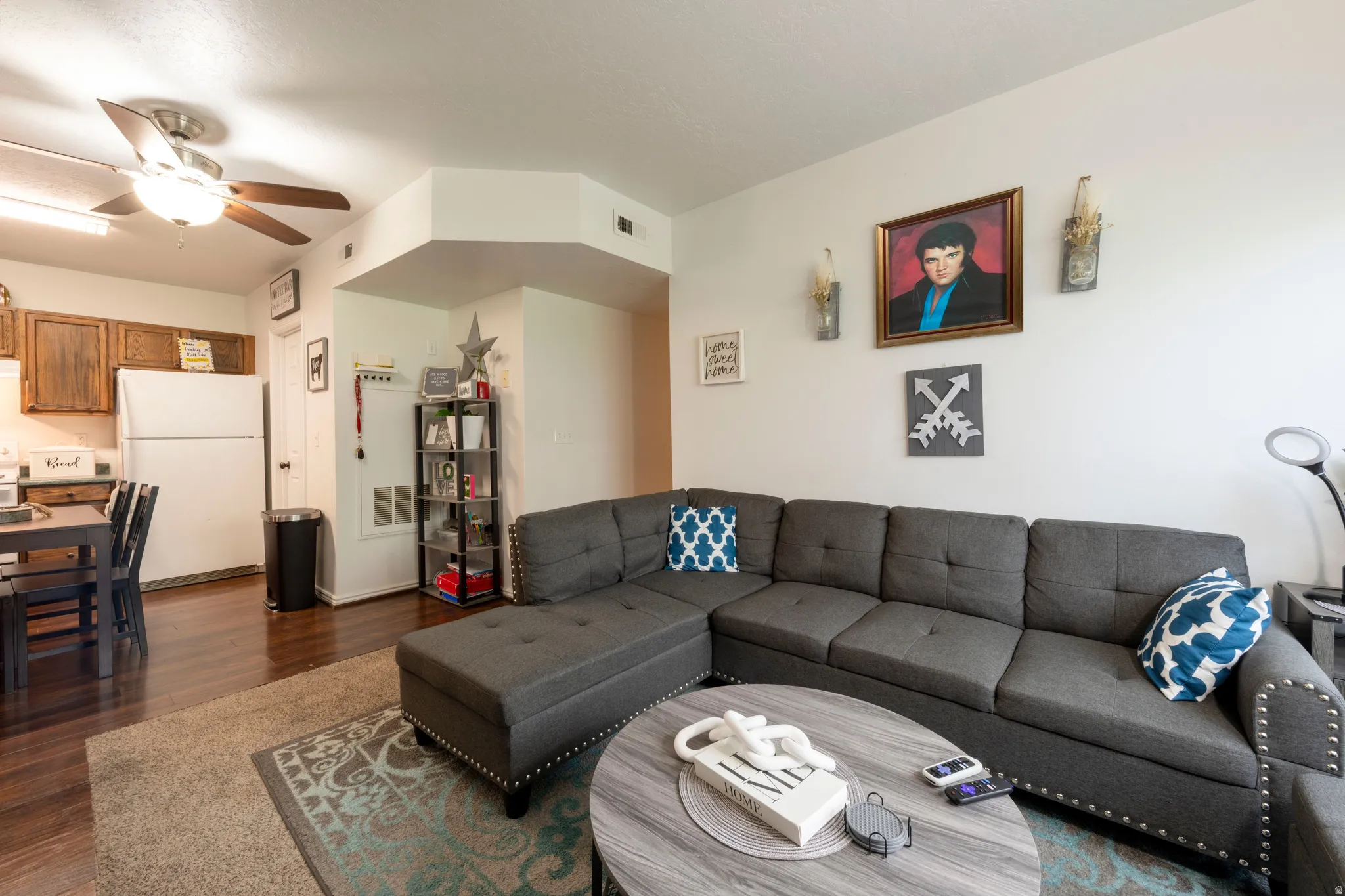 Open-concept living area featuring dark wood-finish flooring, a ceiling fan with integrated lighting, and a kitchen area with wood cabinetry and a white refrigerator