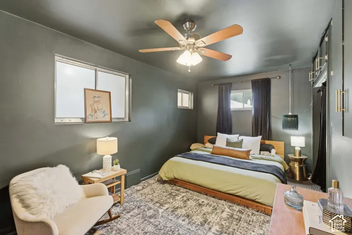Bedroom featuring a ceiling fan with integrated lighting, multiple windows providing natural light, a built-in wall unit with cabinet doors.