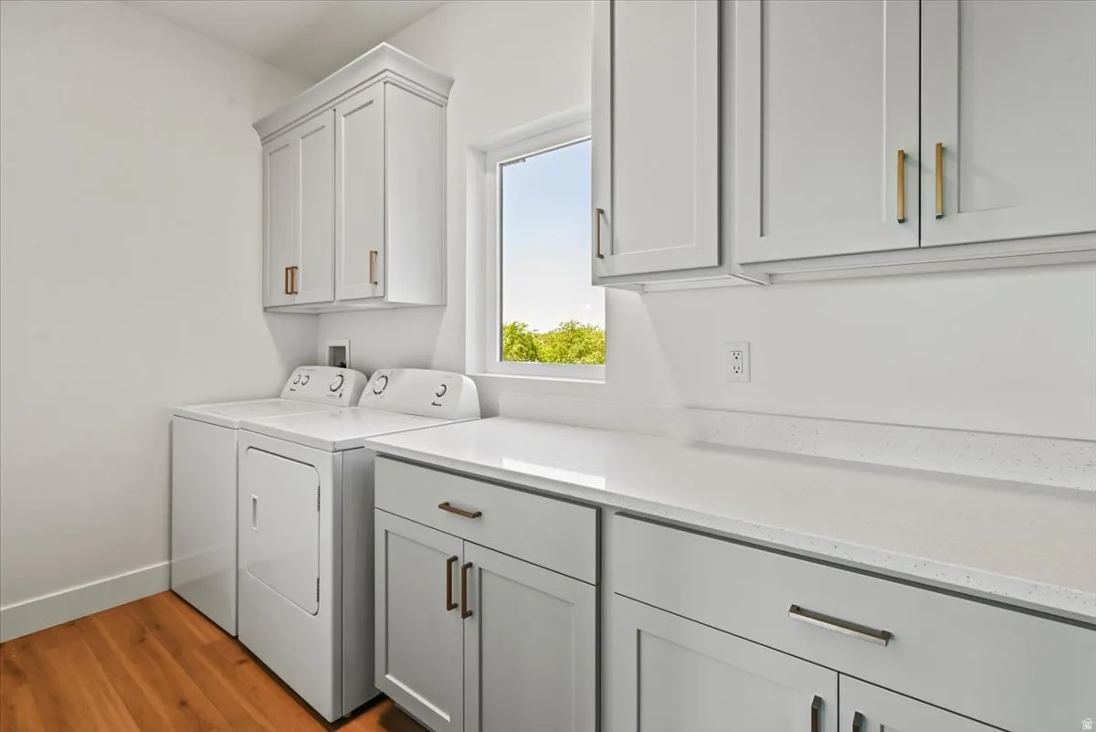 Bright and efficient laundry room featuring white shaker-style cabinetry with brass hardware, a front-load washer and dryer pair, quartz countertops, and abundant storage with overhead cabinets