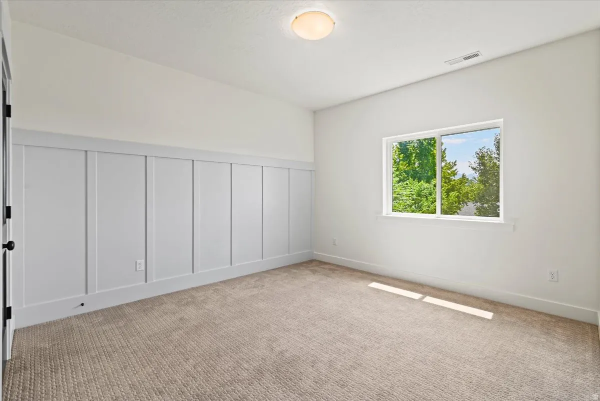 Spacious bedroom featuring built-in closet cabinetry with vertical paneling and hardware pulls, neutral carpet flooring, and abundant natural light from a large window framing mature landscaping and trees