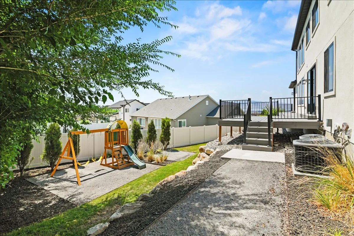 Expansive backyard oasis featuring a multi-level composite deck with black metal railings, mature landscaping with ornamental grasses, a wooden play structure with slide, pea gravel pathways, and manicured lawn space