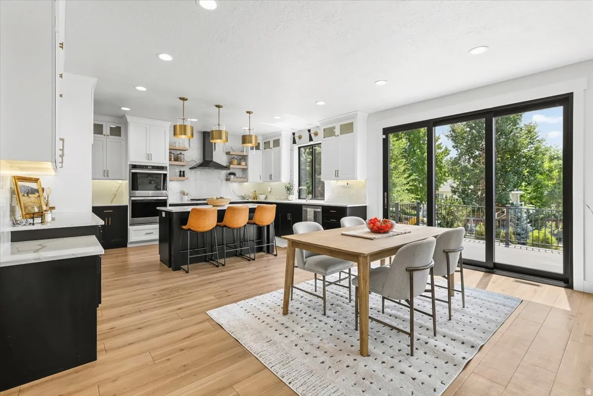 Open-concept kitchen and dining area featuring white cabinetry with brass pendant lighting, a black kitchen island with seating for multiple, wood-finish flooring throughout, and large black-framed sliding glass doors opening to outdoor space