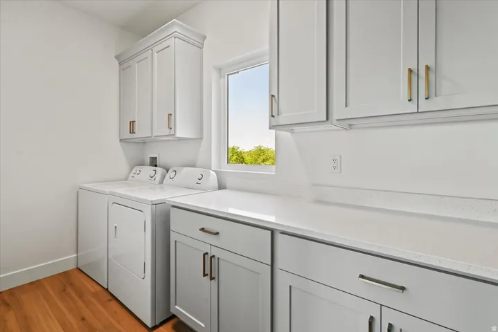 Bright and efficient laundry room featuring white shaker-style cabinetry with brass hardware, a front-load washer and dryer pair, quartz countertops, and abundant storage with overhead cabinets
