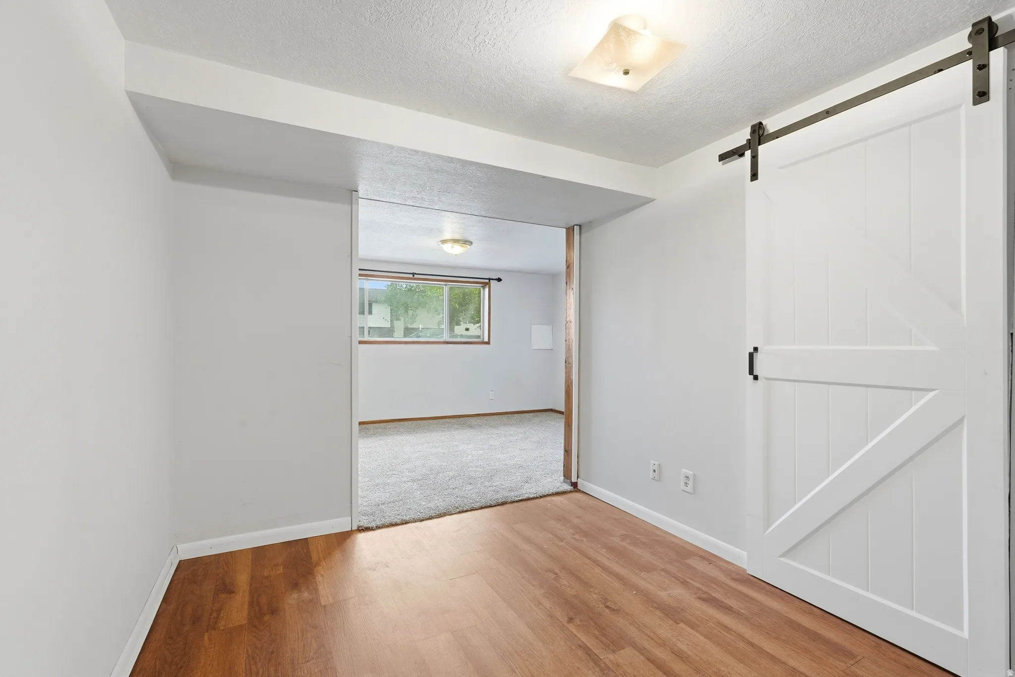 Wood-finish flooring leading into a carpeted room with a barn door and a window