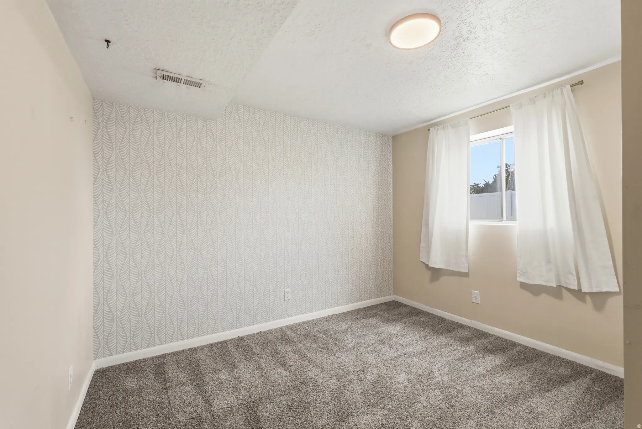 Carpeted room with a patterned accent wall, a single window, and an overhead flush-mount light fixture